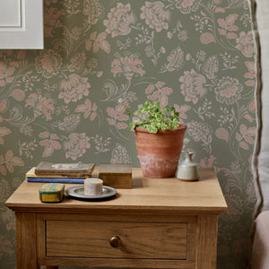 Amelie wallpaper by Curate Space in lichen green, vintage chintz florals in a room with a wooden bedside table and a plant