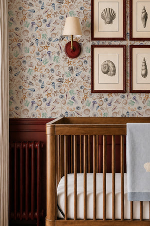 Charlotte wallpaper by Curate Space in light cream, hand-painted seashells and starfish in a nursery with a wooden cot and framed prints