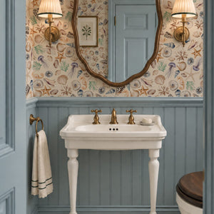 Charlotte Curate Space wallpaper in light cream, hand-painted seashells and starfish in a powder room with a pedestal basin and blue panelling