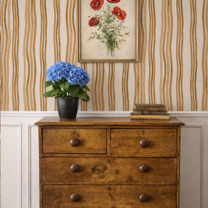 Mara wallpaper by Curate Space in ochre yellow, hand-painted wavy stripes behind a wooden chest of drawers and framed botanical print
