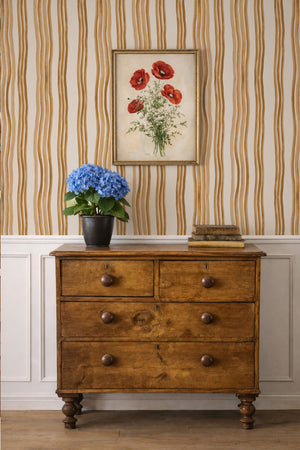 Mara wallpaper by Curate Space in ochre yellow, hand-painted wavy stripes behind a wooden chest of drawers and framed botanical print