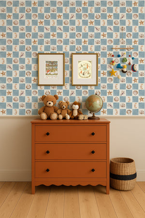 Molly wallpaper by Curate Space in blue, seashells and seahorses on a check behind an orange chest of drawers with framed prints and teddy bears