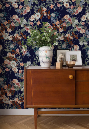 Orla wallpaper by Curate Space in midnight blue, layered florals and foliage over a trellis behind a mid-century sideboard with a vase of foliage